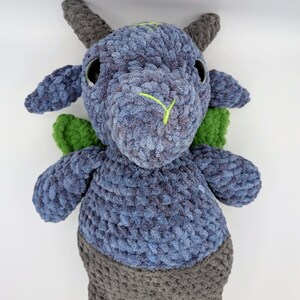 May include: A handmade plush toy in the shape of a mythical creature. The toy is primarily blue with gray accents on the horns, legs, and lower body. It has green wings and black eyes. The face features a neon green stitched design.