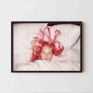 Può includere: Una stampa incorniciata con una persona con capelli rosa acceso sdraiata su un letto bianco. La persona sorride e l'immagine è racchiusa in una cornice nera. La biancheria da letto è bianca.
