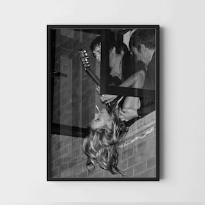 Könnte beinhalten: Schwarz-weiß gerahmtes Foto mit einer Frau mit langen Haaren, einer Zigarette und einem Mann, der sie hält. Ein weiterer Mann ist mit einer Gitarre zu sehen. Das Bild ist vor einer Backsteinmauer.