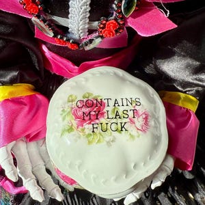 May include: A Day of the Dead figurine with a painted skull face, wearing a black dress with pink accents and a floral headdress. The figurine holds a white, round container with the text "CONTAINS MY LAST FUCK".