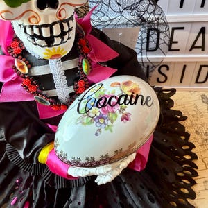 May include: A Day of the Dead figurine with a decorated skull, floral necklace, and black dress. It holds a white egg-shaped container with "Cocaine" and floral designs. The background includes a sign that says "TAKE DEAD SIDE".