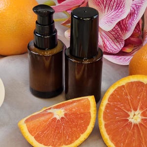 May include: Two brown glass bottles with black pumps and caps are displayed with orange slices and whole oranges. The background includes white and pink orchid flowers. The image has a natural, fresh aesthetic.