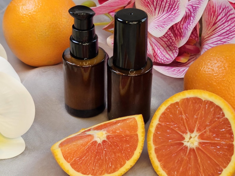 May include: Two brown glass bottles with black pumps and caps are displayed with orange slices and whole oranges. The background includes white and pink orchid flowers. The image has a natural, fresh aesthetic.