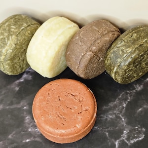 May include: Five solid shampoo bars in different colors: green, white, brown, green, and brown. The bars are arranged in a row on a black marble surface.