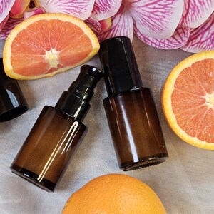 May include: Two amber glass pump bottles with black pumps and caps are displayed alongside orange and grapefruit slices and pink and purple orchid blossoms. The bottles are empty and ready to be filled with product.