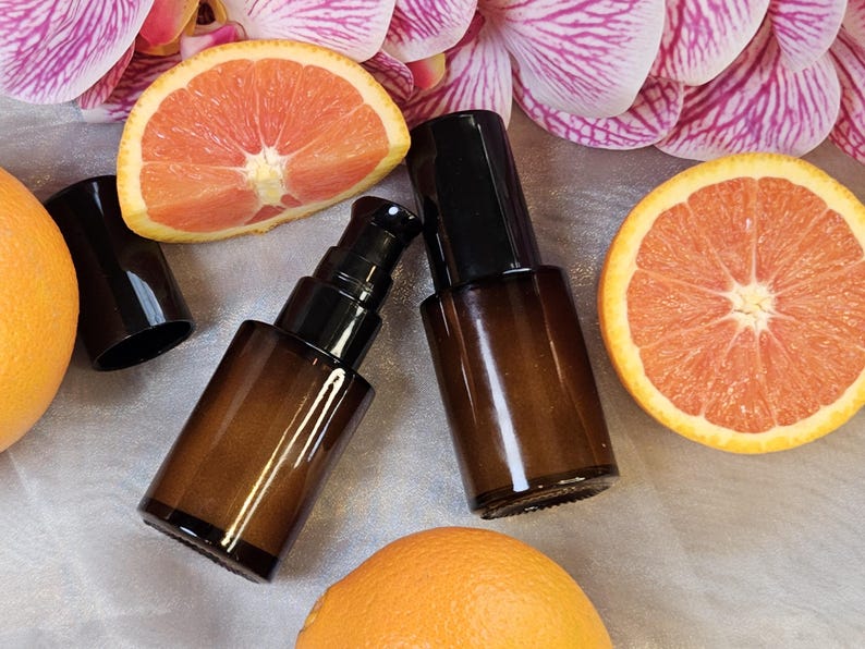 May include: Two amber glass pump bottles with black pumps and caps are displayed alongside orange and grapefruit slices and pink and purple orchid blossoms. The bottles are empty and ready to be filled with product.