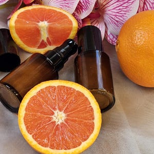 May include: Brown glass bottles with black caps and pump dispensers are arranged with sliced and whole oranges. The oranges are bright orange with a reddish interior. Pink and white flowers are in the background.
