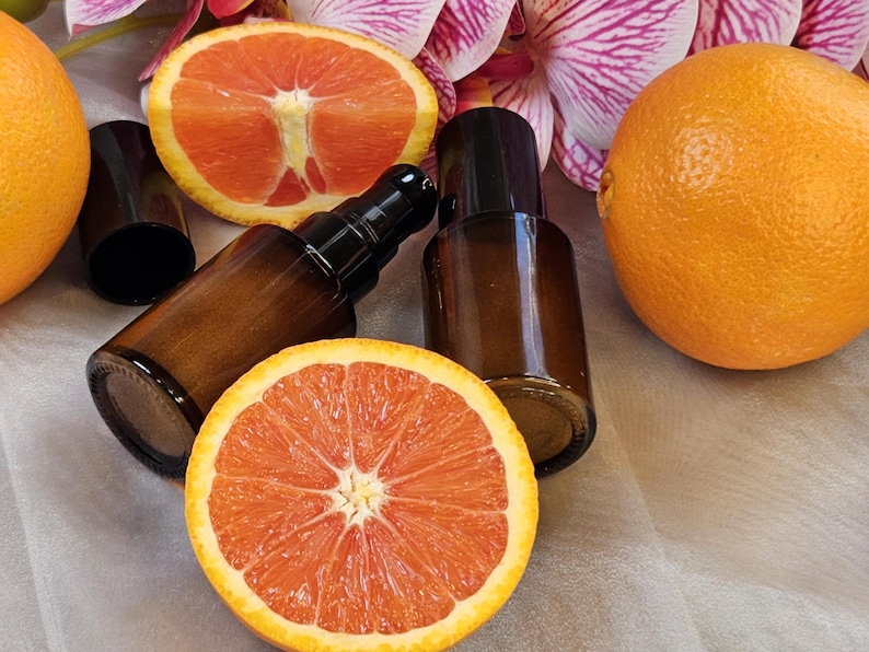 May include: Brown glass bottles with black caps and pump dispensers are arranged with sliced and whole oranges. The oranges are bright orange with a reddish interior. Pink and white flowers are in the background.