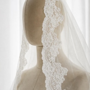 May include: A white wedding veil featuring intricate floral lace trim along the edges. The sheer, lightweight fabric drapes over a mannequin head and shoulders, showcasing the detailed scalloped lace pattern that frames the face.