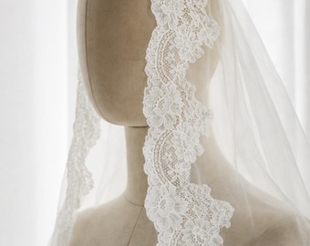 May include: A white wedding veil featuring intricate floral lace trim along the edges. The sheer, lightweight fabric drapes over a mannequin head and shoulders, showcasing the detailed scalloped lace pattern that frames the face.