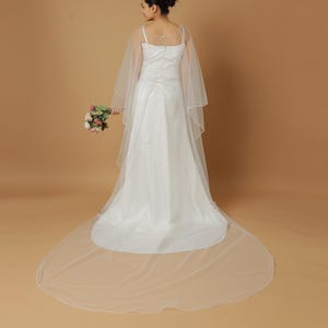 A bride wearing an elegant lace wedding gown is adorned with a sheer, white tulle bridal cape. The cape features a soft round neckline and flows gracefully over her shoulders, ending just above the waist in front and trailing longer in the back. The fitted gown has a subtle floral texture and a strapless sweetheart neckline. The setting is softly lit with neutral tones, featuring white curtains and minimal décor, highlighting the romantic and refined look.