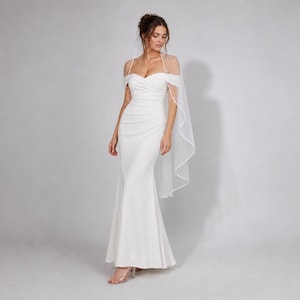 May include: A floor-length white gown featuring a sweetheart neckline, off-the-shoulder straps, and a ruched bodice that flows into a fitted skirt. A sheer, floor-length veil with a thin white trim is draped over the shoulders and back.