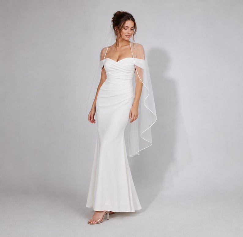 May include: A floor-length white gown featuring a sweetheart neckline, off-the-shoulder straps, and a ruched bodice that flows into a fitted skirt. A sheer, floor-length veil with a thin white trim is draped over the shoulders and back.