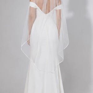 May include: A white wedding veil made of sheer tulle fabric featuring a delicate satin ribbon trim along the edges. The veil is attached to a hair comb and drapes down the back, falling to a mid-length point, complementing a floor-length gown.