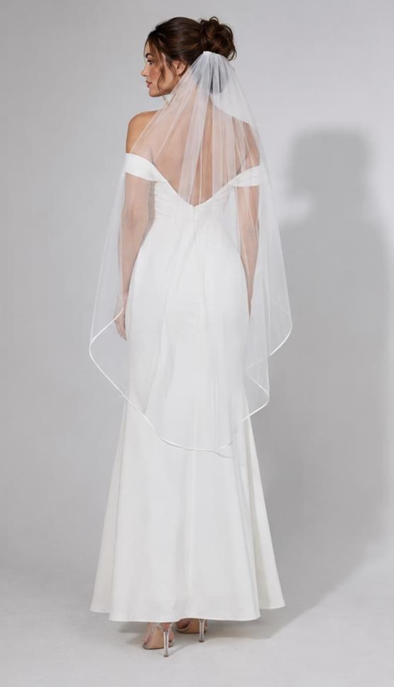 May include: A white wedding veil made of sheer tulle fabric featuring a delicate satin ribbon trim along the edges. The veil is attached to a hair comb and drapes down the back, falling to a mid-length point, complementing a floor-length gown.