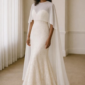 Beautiful bride in a modern white chiffon evening cape dress with a flowing sheer overlay and elegant satin base, standing gracefully against a soft beige background with warm studio lights highlighting her natural beauty. The chiffon cape adds ethereal movement, perfect for bridal photoshoots, rehearsal dinners, or wedding receptions. A minimalist yet luxurious fashion choice for the modern bride seeking timeless elegance.