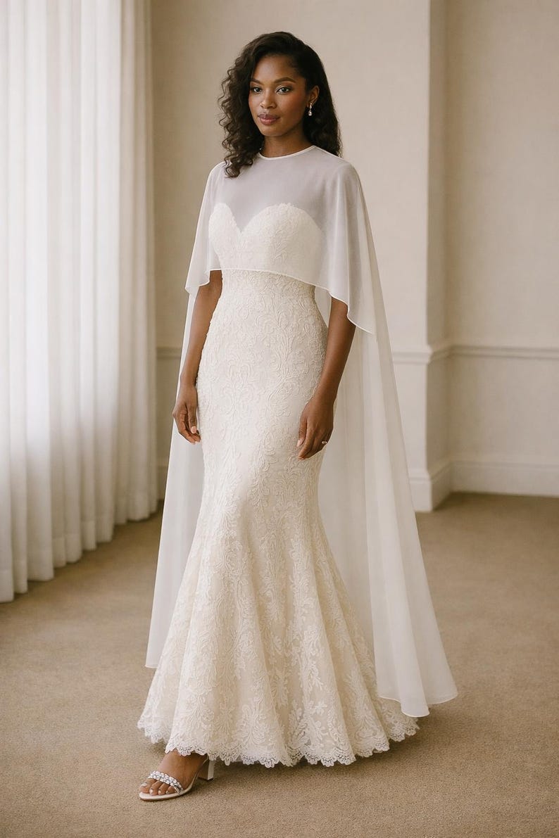 Beautiful bride in a modern white chiffon evening cape dress with a flowing sheer overlay and elegant satin base, standing gracefully against a soft beige background with warm studio lights highlighting her natural beauty. The chiffon cape adds ethereal movement, perfect for bridal photoshoots, rehearsal dinners, or wedding receptions. A minimalist yet luxurious fashion choice for the modern bride seeking timeless elegance.