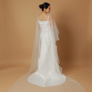 A bride wearing an elegant lace wedding gown is adorned with a sheer, white tulle bridal cape. The cape features a soft round neckline and flows gracefully over her shoulders, ending just above the waist in front and trailing longer in the back. The fitted gown has a subtle floral texture and a strapless sweetheart neckline. The setting is softly lit with neutral tones, featuring white curtains and minimal décor, highlighting the romantic and refined look.