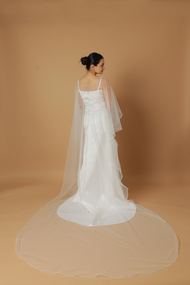 A bride wearing an elegant lace wedding gown is adorned with a sheer, white tulle bridal cape. The cape features a soft round neckline and flows gracefully over her shoulders, ending just above the waist in front and trailing longer in the back. The fitted gown has a subtle floral texture and a strapless sweetheart neckline. The setting is softly lit with neutral tones, featuring white curtains and minimal décor, highlighting the romantic and refined look.