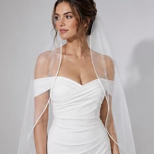 May include: A white wedding veil featuring a delicate satin ribbon trim along the edge, draped over a white off-the-shoulder dress with a sweetheart neckline and ruched bodice detailing that creates a fitted silhouette across the torso.