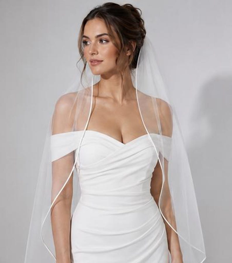 May include: A white wedding veil featuring a delicate satin ribbon trim along the edge, draped over a white off-the-shoulder dress with a sweetheart neckline and ruched bodice detailing that creates a fitted silhouette across the torso.