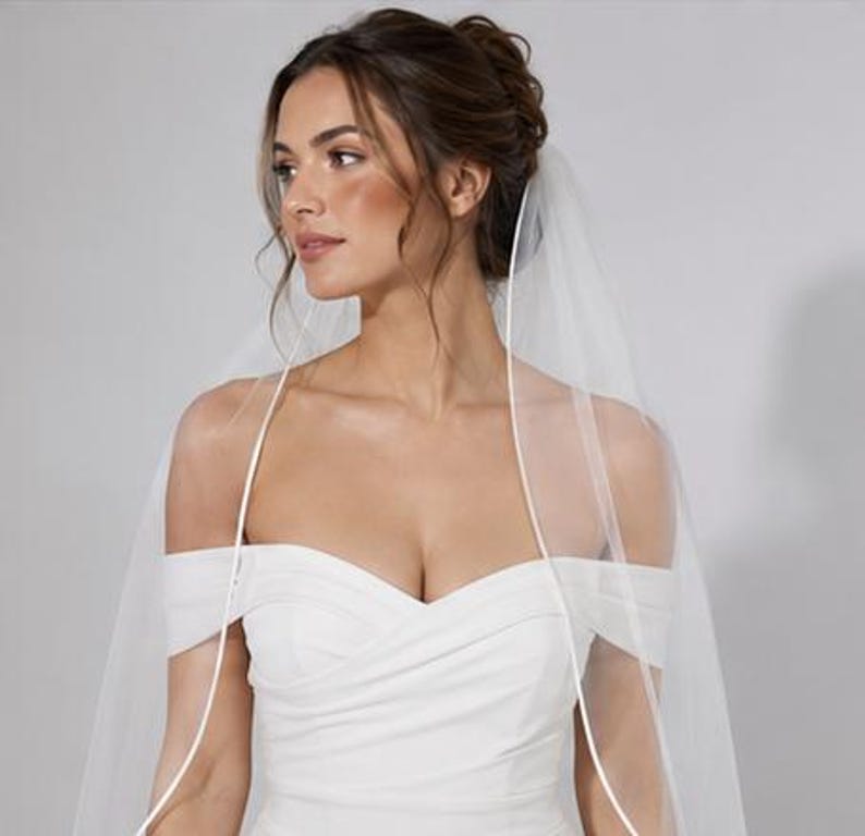 May include: A white off-the-shoulder wedding dress featuring a sweetheart neckline with draped fabric across the bodice. A sheer, floor-length white veil with a thin satin ribbon trim is draped over the head and shoulders, framing the upper body.
