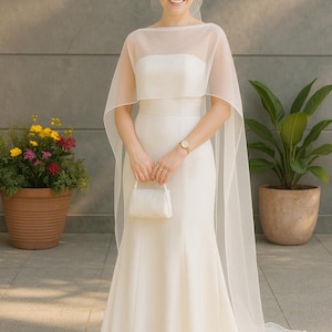 May include: A white bridal gown with a sheer cape and a small white handbag. The dress features a fitted bodice and a mermaid skirt. The bride wears a gold watch. Potted plants and flowers are in the background.
