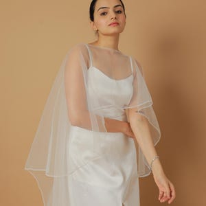 A bride wearing an elegant lace wedding gown is adorned with a sheer, white tulle bridal cape. The cape features a soft round neckline and flows gracefully over her shoulders, ending just above the waist in front and trailing longer in the back. The fitted gown has a subtle floral texture and a strapless sweetheart neckline. The setting is softly lit with neutral tones, featuring white curtains and minimal décor, highlighting the romantic and refined look.