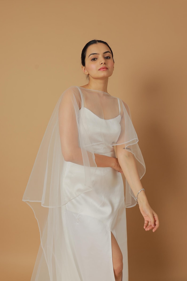 A bride wearing an elegant lace wedding gown is adorned with a sheer, white tulle bridal cape. The cape features a soft round neckline and flows gracefully over her shoulders, ending just above the waist in front and trailing longer in the back. The fitted gown has a subtle floral texture and a strapless sweetheart neckline. The setting is softly lit with neutral tones, featuring white curtains and minimal décor, highlighting the romantic and refined look.