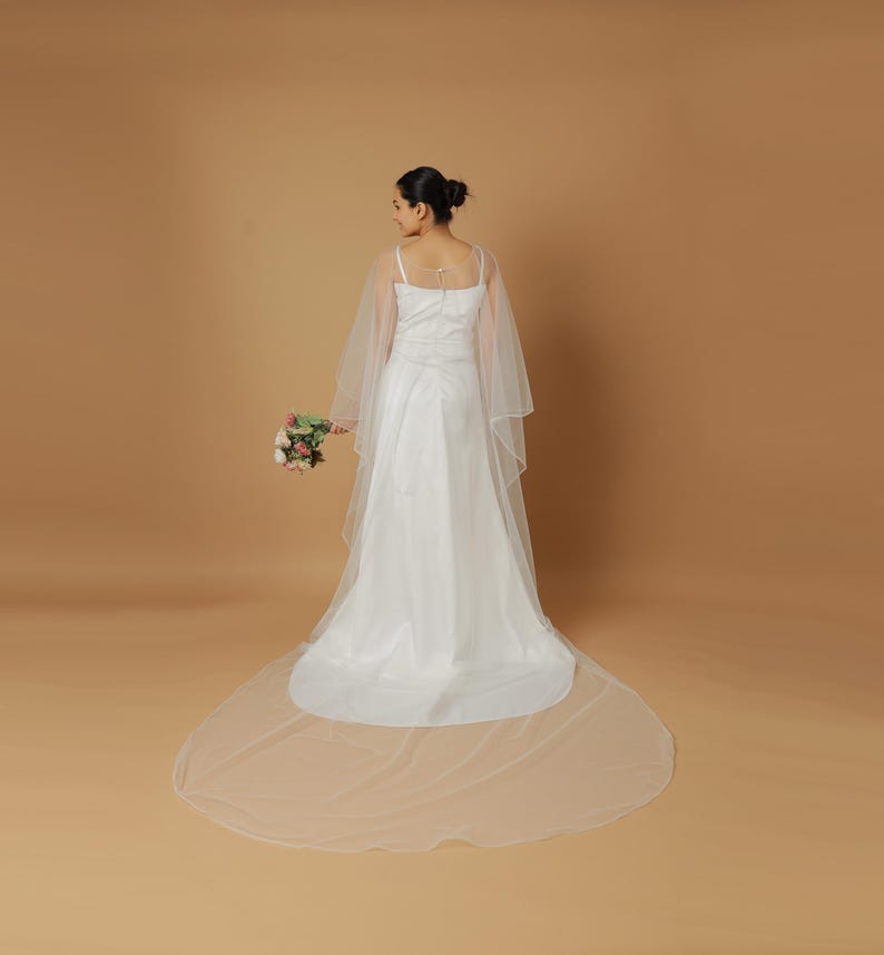 A bride wearing an elegant lace wedding gown is adorned with a sheer, white tulle bridal cape. The cape features a soft round neckline and flows gracefully over her shoulders, ending just above the waist in front and trailing longer in the back. The fitted gown has a subtle floral texture and a strapless sweetheart neckline. The setting is softly lit with neutral tones, featuring white curtains and minimal décor, highlighting the romantic and refined look.