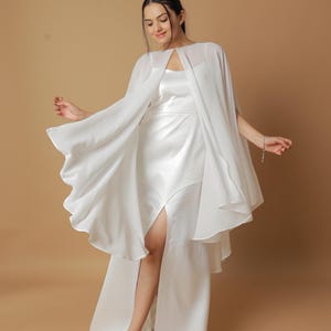 May include: A white satin gown with a high slit, paired with a flowing chiffon cape. The cape has a keyhole neckline and a scalloped hem. The dress is a classic bridal or formal wear style.