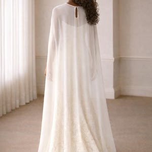 Beautiful bride in a modern white chiffon evening cape dress with a flowing sheer overlay and elegant satin base, standing gracefully against a soft beige background with warm studio lights highlighting her natural beauty. The chiffon cape adds ethereal movement, perfect for bridal photoshoots, rehearsal dinners, or wedding receptions. A minimalist yet luxurious fashion choice for the modern bride seeking timeless elegance.