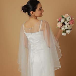 A bride wearing an elegant lace wedding gown is adorned with a sheer, white tulle bridal cape. The cape features a soft round neckline and flows gracefully over her shoulders, ending just above the waist in front and trailing longer in the back. The fitted gown has a subtle floral texture and a strapless sweetheart neckline. The setting is softly lit with neutral tones, featuring white curtains and minimal décor, highlighting the romantic and refined look.