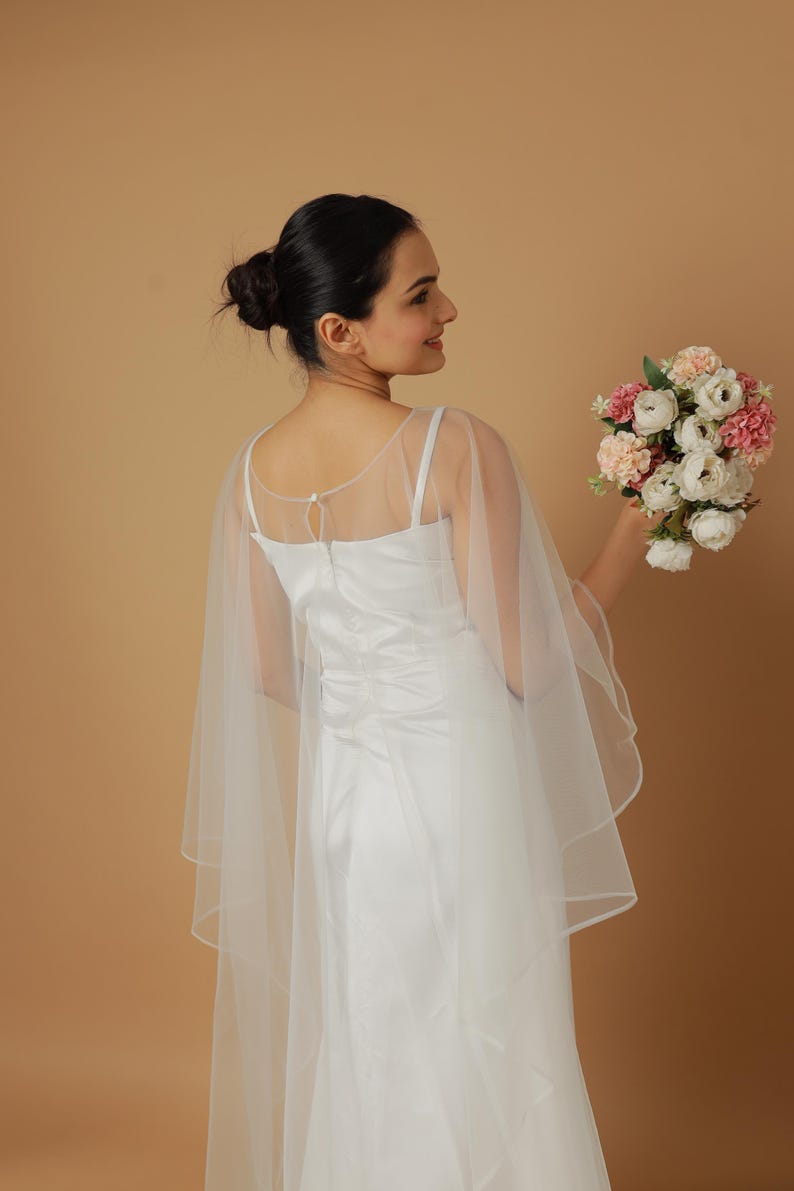 A bride wearing an elegant lace wedding gown is adorned with a sheer, white tulle bridal cape. The cape features a soft round neckline and flows gracefully over her shoulders, ending just above the waist in front and trailing longer in the back. The fitted gown has a subtle floral texture and a strapless sweetheart neckline. The setting is softly lit with neutral tones, featuring white curtains and minimal décor, highlighting the romantic and refined look.