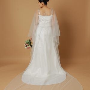 A bride wearing an elegant lace wedding gown is adorned with a sheer, white tulle bridal cape. The cape features a soft round neckline and flows gracefully over her shoulders, ending just above the waist in front and trailing longer in the back. The fitted gown has a subtle floral texture and a strapless sweetheart neckline. The setting is softly lit with neutral tones, featuring white curtains and minimal décor, highlighting the romantic and refined look.