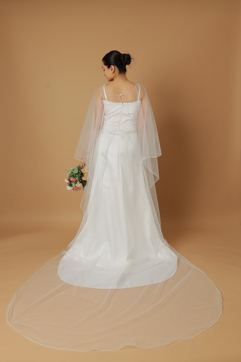 A bride wearing an elegant lace wedding gown is adorned with a sheer, white tulle bridal cape. The cape features a soft round neckline and flows gracefully over her shoulders, ending just above the waist in front and trailing longer in the back. The fitted gown has a subtle floral texture and a strapless sweetheart neckline. The setting is softly lit with neutral tones, featuring white curtains and minimal décor, highlighting the romantic and refined look.