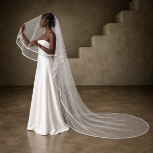 May include: A floor-length white wedding veil made of sheer tulle fabric featuring a smooth satin ribbon trim along the entire edge. The veil is attached to a hair comb and drapes down to the floor, creating a long, flowing train behind the wearer.