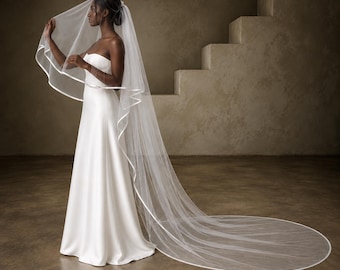May include: A floor-length white wedding veil made of sheer tulle fabric featuring a smooth satin ribbon trim along the entire edge. The veil is attached to a hair comb and drapes down to the floor, creating a long, flowing train behind the wearer.