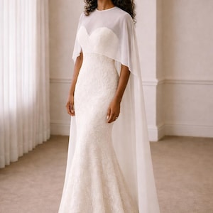 Beautiful bride in a modern white chiffon evening cape dress with a flowing sheer overlay and elegant satin base, standing gracefully against a soft beige background with warm studio lights highlighting her natural beauty. The chiffon cape adds ethereal movement, perfect for bridal photoshoots, rehearsal dinners, or wedding receptions. A minimalist yet luxurious fashion choice for the modern bride seeking timeless elegance.