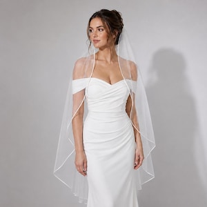 May include: A white floor-length wedding dress featuring an off-the-shoulder neckline with ruched fabric across the bodice and a fitted silhouette. A sheer, lightweight white veil with a thin satin ribbon trim drapes over the shoulders.