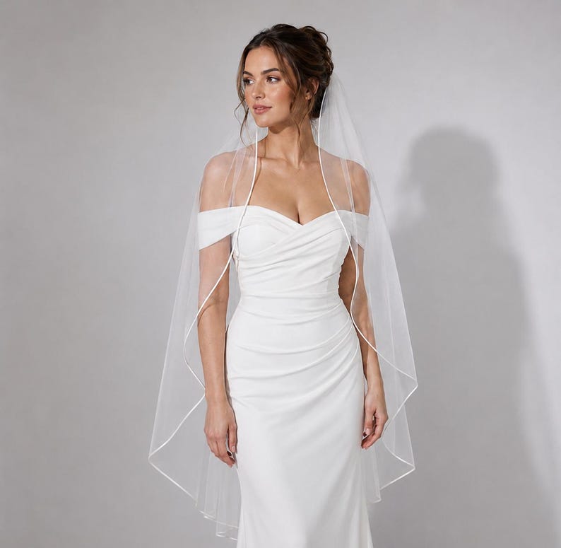 May include: A white floor-length wedding dress featuring an off-the-shoulder neckline with ruched fabric across the bodice and a fitted silhouette. A sheer, lightweight white veil with a thin satin ribbon trim drapes over the shoulders.