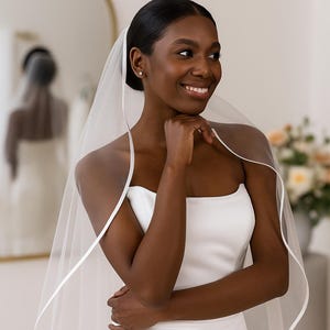 Close-up image of a dark-skinned bridal model smiling warmly while wearing a satin edge veil. The soft beige background highlights her radiant expression and the elegant detailing of the veil.