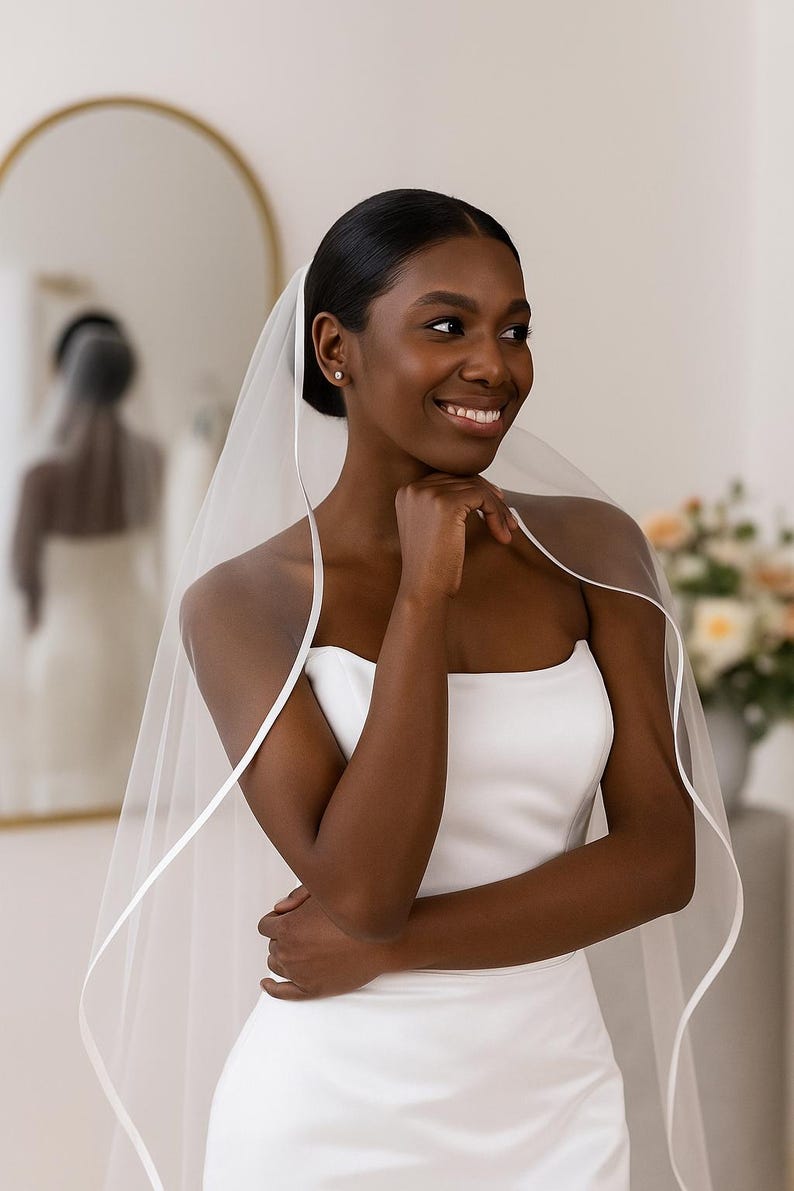 Close-up image of a dark-skinned bridal model smiling warmly while wearing a satin edge veil. The soft beige background highlights her radiant expression and the elegant detailing of the veil.