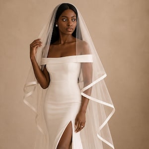 May include: A floor-length white wedding dress featuring an off-the-shoulder neckline and a high thigh slit. The dress is paired with a long, sheer white veil that has a solid white ribbon trim along the edges, draping gracefully over the shoulders.