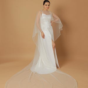 A bride wearing an elegant lace wedding gown is adorned with a sheer, white tulle bridal cape. The cape features a soft round neckline and flows gracefully over her shoulders, ending just above the waist in front and trailing longer in the back. The fitted gown has a subtle floral texture and a strapless sweetheart neckline. The setting is softly lit with neutral tones, featuring white curtains and minimal décor, highlighting the romantic and refined look.