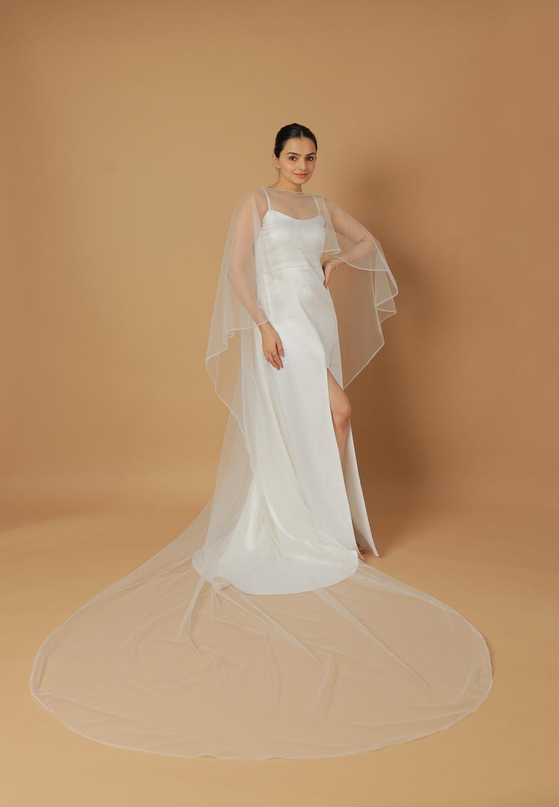 A bride wearing an elegant lace wedding gown is adorned with a sheer, white tulle bridal cape. The cape features a soft round neckline and flows gracefully over her shoulders, ending just above the waist in front and trailing longer in the back. The fitted gown has a subtle floral texture and a strapless sweetheart neckline. The setting is softly lit with neutral tones, featuring white curtains and minimal décor, highlighting the romantic and refined look.