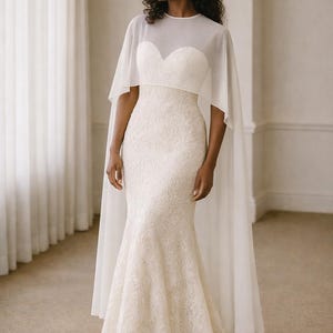 Beautiful bride in a modern white chiffon evening cape dress with a flowing sheer overlay and elegant satin base, standing gracefully against a soft beige background with warm studio lights highlighting her natural beauty. The chiffon cape adds ethereal movement, perfect for bridal photoshoots, rehearsal dinners, or wedding receptions. A minimalist yet luxurious fashion choice for the modern bride seeking timeless elegance.