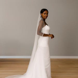 side view image of a dark-skinned bridal model while wearing a Ivory tulle satin edge veil. The soft beige background highlights her radiant expression and the elegant detailing of the veil.