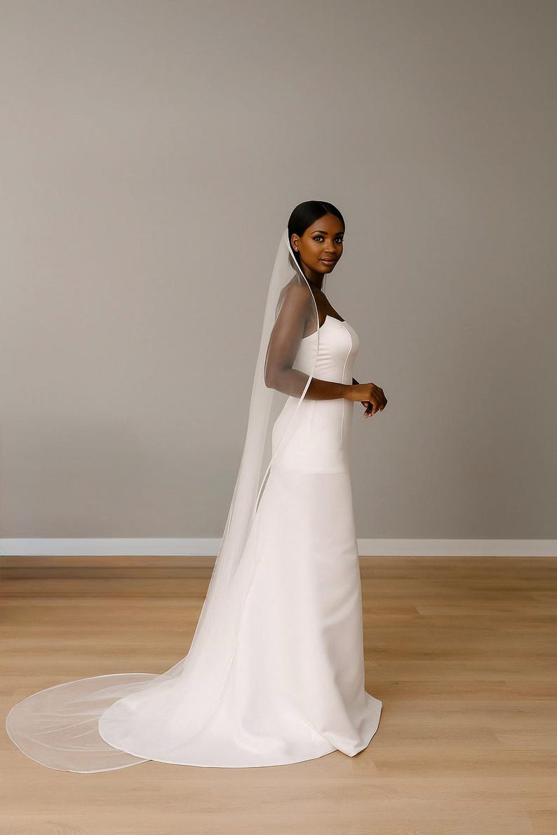 side view image of a dark-skinned bridal model while wearing a Ivory tulle satin edge veil. The soft beige background highlights her radiant expression and the elegant detailing of the veil.