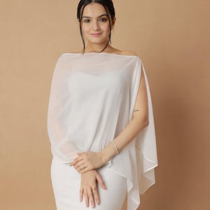 Può includere: Un top bianco con mantella a spalle scoperte e un vestito abbinato. Il top con mantella ha un design fluido e trasparente, e l'abito è aderente. L'outfit è interamente bianco, creando un look pulito ed elegante. Lo sfondo è di colore beige.